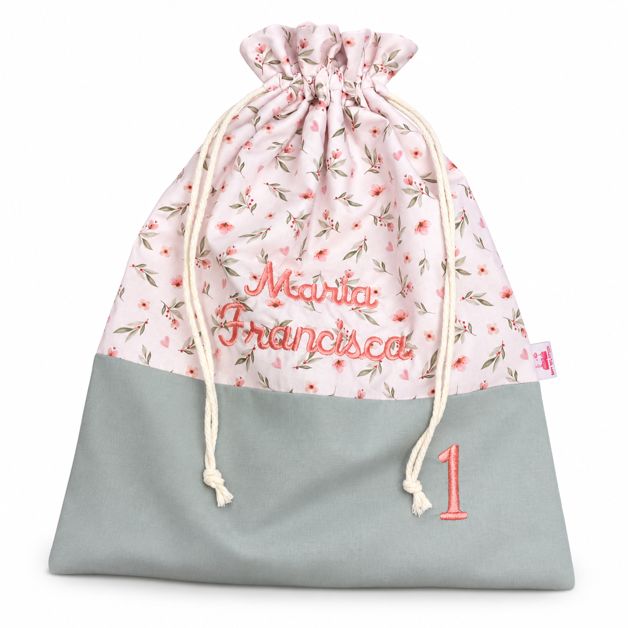 Saco de tecido rosa com flores, cordões trançados e bordado “Maria Francisca” em vermelho.